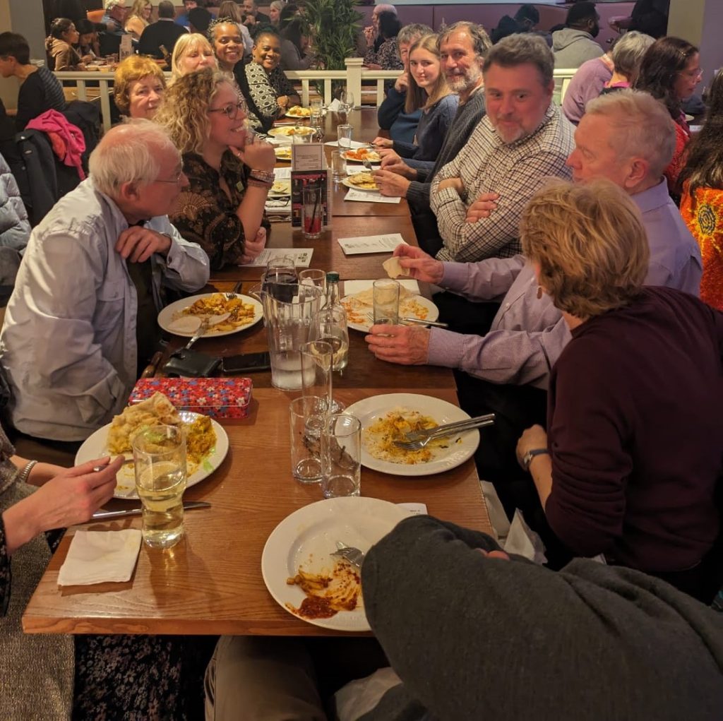 Photograph of the last Vaughan supper. People are seated around a long table. Some of talking intensively others are looking at the camera.