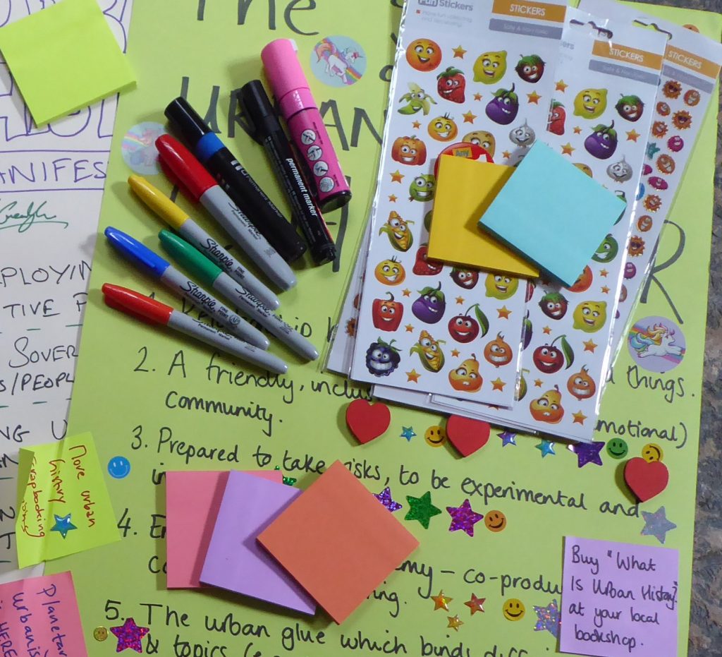 Image of stickers, marker pens and sticky notes on a background of hand-written posters with text on them. The text on the sticky notes and posters are largely concealed by the things on top.