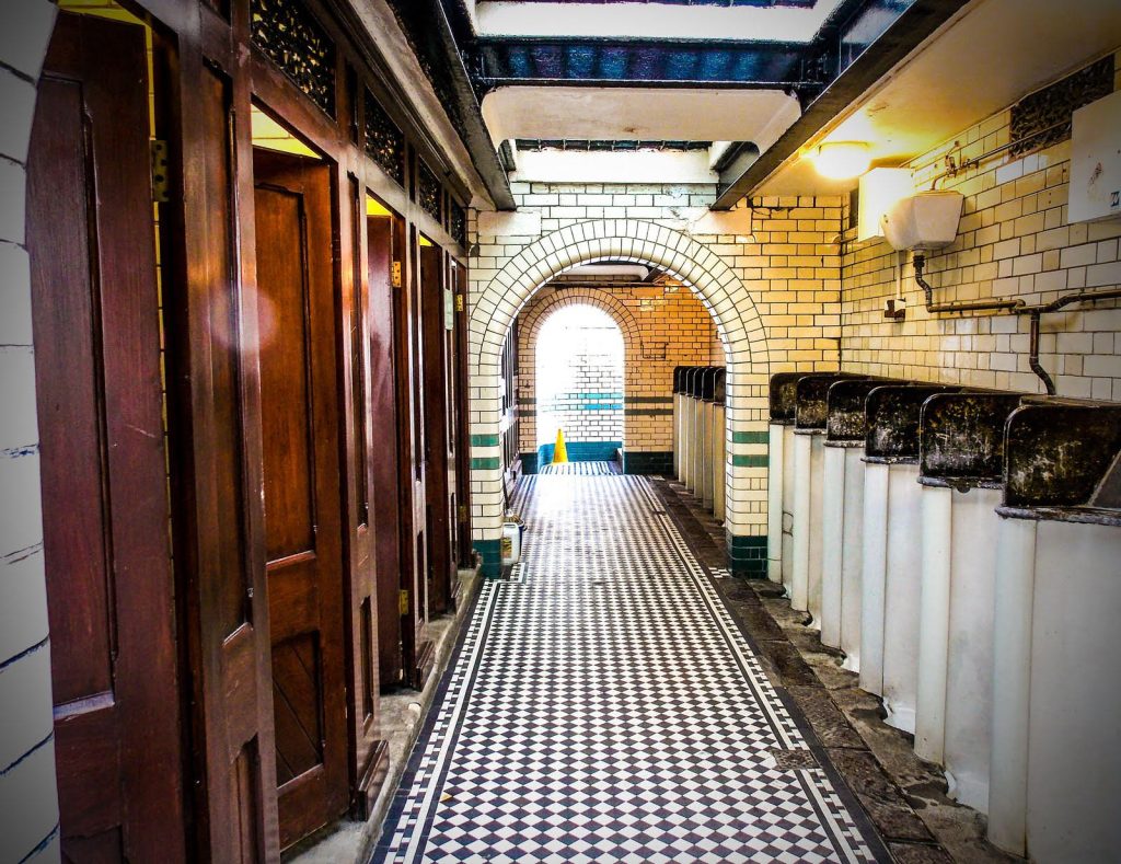 The interior of a men's public toilet. On the right there is a row of urinals in waist height booths, covered in white tiles. On the left at the wooden doors to WCs. The floor between is of black and white tiles and leads through tow arches.