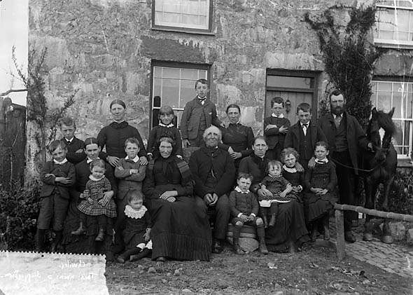 An extended family group of 19 people across three generations. The family is sitting outside their cottage in Wales, UK. The image is from the National Library of Wales via Wikimedia commons.