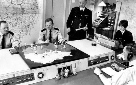 Black and white image of a police control room. There are maps on the walls and three policeman and one police woman sit around the control table which has various gadgets. Another officer is standing.
